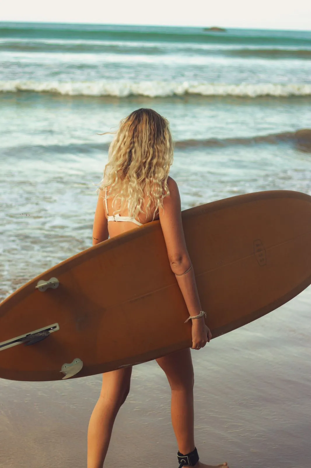 Young girl carrying a surfboard at Unawatuna Beach in Sri Lanka, walking along the sandy shore with waves crashing in the background and palm trees swaying in the breeze.